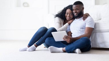 Bir çift, tablet bir cihaz kullanarak halı kaplı bir zeminde oturuyor. Kadın başını erkeğin omzuna yaslıyor ve mutlu ve rahat görünüyorlar..