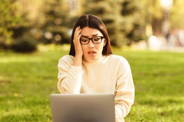 Krem rengi bir kazak giymiş Asyalı genç bir kadın güneşli bir parkta çimlerin üzerinde oturuyor. Dizüstü bilgisayarına bakarken şaşırmış ve paniğe kapılmış bir halde bakıyor..