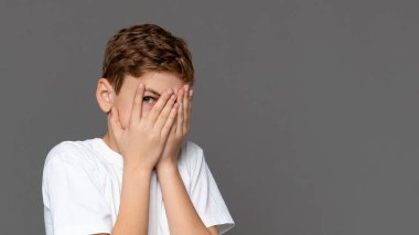 Beyaz tişörtlü, tarafsız bir çocuk elleriyle yüzünü kapatıyor. Utanç, korku ya da utangaçlık ifade ediyor.