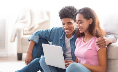 Afro-Amerikalı genç çift oturma odasında oturmuş dizüstü bilgisayara bakıyor. Adamın kolu kızın omzunda ve ikisi de laptopta gördüklerine odaklanmış..
