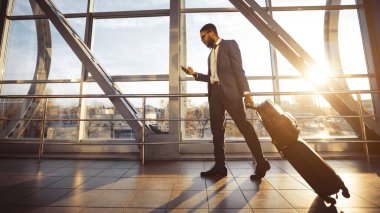 Afro-Amerikalı bir adam havaalanı terminalinde telefonunu kullanıyor, bavulunun yanında duruyor. Ortam modern ve hareketli, etkin bir yolculuk, güneş parlaması...