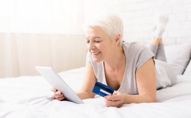 Online bankacılık. Modern kıdemli kadın, ev, boş alan yatakta yatarken, dijital tablet üzerinden onu kredi kartı dengesini kontrol