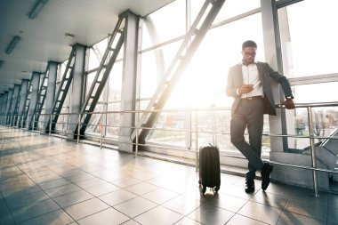 Afrika kökenli Amerikalı işadamı modern bir havaalanında bavuluyla dikilirken telefonunu kullanıyor. Sahne yoğun ve profesyoneldir, seyahat ve iletişimin altını çizmek, güneş parlaması