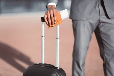 Pasaportu ve uçuş biletleri olan siyah bir işadamı elini bavulun üzerinde tutarak havaalanının dışında poz veriyor.