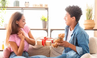 Afro-Amerikalı bir genç kız arkadaşına hediyesiyle ve oyuncak ayıyla oturma odasındaki koltukta sürpriz yapıyor. Kız sevinçle gülümseyip ellerini çırparken hediyeleri elinde tutuyor..