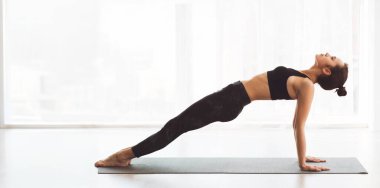 Genç bir kadın, yoga minderindeki ters tahta duruşu ile çekirdek gücünü ve dengesini arttırıyor. Geniş, güneşli stüdyo, sabah egzersizi için huzurlu bir ortam yaratıyor.