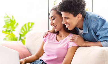 Siyah çift aydınlık bir odada beyaz bir kanepede oturuyor. Kız kanepenin sol tarafında dizüstü bilgisayar kullanırken, adam arkasından eğiliyor, gülümsüyor ve ona bakıyor.