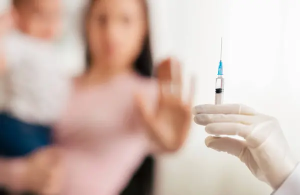 stock image A parent holding a young child raises their hand in refusal as a healthcare worker attempts to administer a vaccination in a clinic setting.