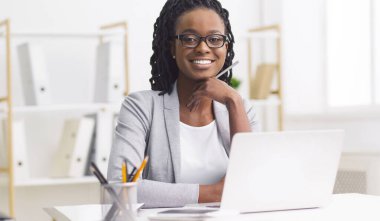 Kendini işine adamış profesyonel siyahi bir kadın masasında dizüstü bilgisayarla çalışıyor. Ofis modern ve düzenli, bağlılığını yansıtıyor ve görevlerine odaklanıyor..