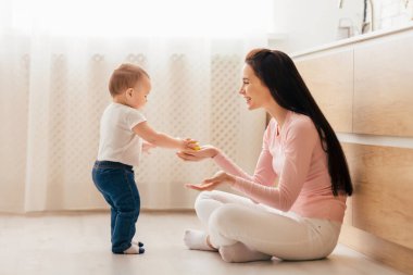 Bir anne ve yeni yürümeye başlayan çocuğu, aydınlık ve modern bir mutfakta birlikte kaliteli zaman geçiriyorlar. Anne yerde oturuyor, gülümsüyor ve önünde duran yavrusunu cesaretlendiriyor.