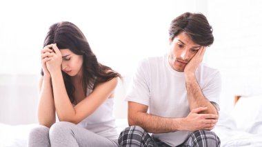 Yüzlerinde hüzünlü ifadelerle beyaz bir yatakta oturan bir çift. Kadın gri bir bluz ve pantolon giyiyor, ve erkek beyaz bir tişört ve ekose pantolon giyiyor..
