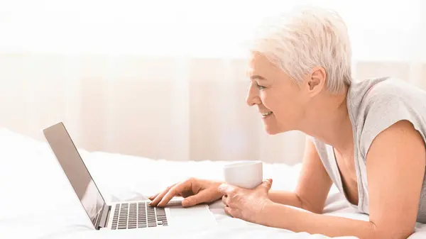 stock image Senior woman is laying on a bed while using a laptop. She appears focused on the screen, with her hands typing or scrolling. The room is dimly lit, creating a cozy atmosphere, side view