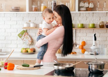 Beyaz gömlek ve kot pantolon giyen omuz hizasında saçlı bir kadın mutfakta durmuş, kucağında bir bebek tutuyor. Bebek mavi bir battaniyeye sarılmış ve merakla kameraya bakıyor..