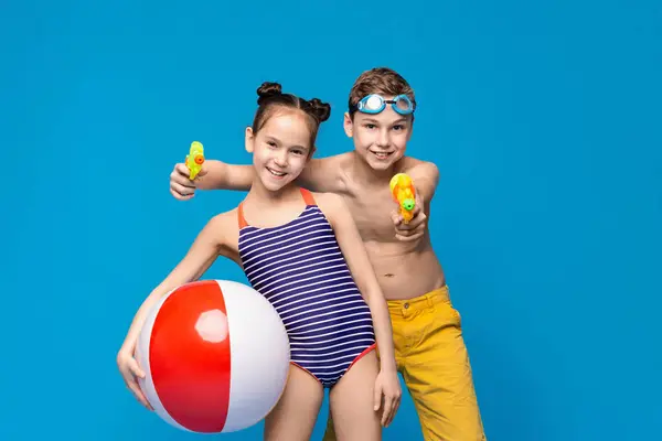 stock image Two happy children are having fun with summer activities. A girl holds a beach ball while a boy aims a water gun. Both are dressed in swimwear, with a vibrant blue backdrop emphasizing lively mood.