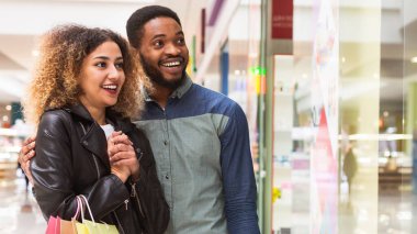 Neşeli bir Afrikalı Amerikalı erkek ve kadın alışveriş merkezinin koridorunda dolaşıyorlar. Yüz ifadeleri, çok hoş şeyler satın aldıklarını gösteriyor.
