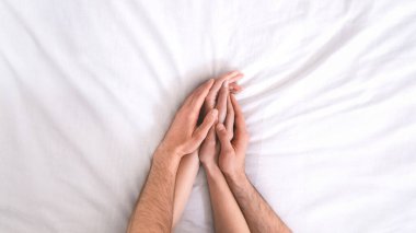 İki elin üst üste yakın çekimi, bir erkek ve bir kadın, beyaz yatak örtüsüne dolanmış. Elleri sevgi dolu ve samimi bir şekilde yerleştirilmiş. Bu da romantik ya da samimi bir bağ olduğunu gösteriyor.
