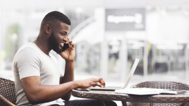 Kafenin dışındaki bir masada oturan bir adam dizüstü bilgisayarıyla çalışıyor ve telefonuyla konuşuyor. Gülümsüyor ve işini seviyor gibi görünüyor..