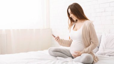 Neşeli hamile kadın akıllı telefondan bakıyor, evde yatakta oturuyor, boş bir alan.