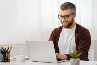 Masada oturan bir adam dizüstü bilgisayarının ekranına odaklanmış. Ekrana dikkatle bakarken elleri klavyeye yazı yazıyor. Çalışma alanı düzenlidir.
