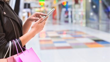Meşgul bir alışveriş alanında kesilen bir kadın diğer eliyle cep telefonunu kullanırken bir elinde birden fazla alışveriş torbası tutuyor. Telefonuna odaklanmış görünüyor, kopyalama alanı.