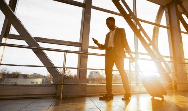 Zenci bir iş gezgini havaalanında yanında bavuluyla telefonuyla konuşuyor. Ortam hareketli ve profesyonel, yoğun bir seyahat günü, güneş parlaması