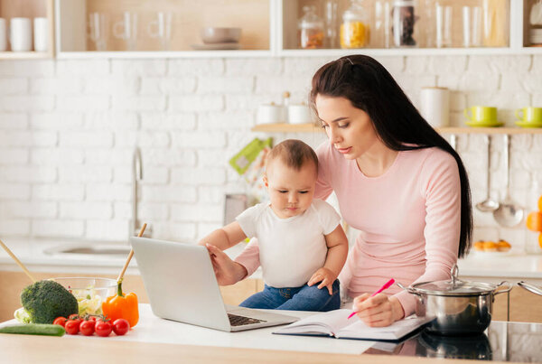 A mother multitasks in a bright, modern kitchen, working on her laptop and taking notes while holding her baby. Fresh vegetables and a pot on the stove are visible in the background.