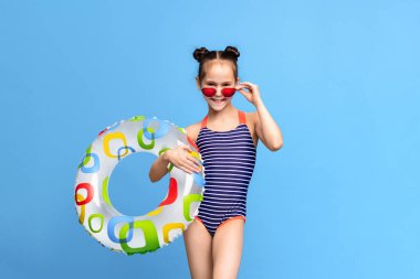 Çizgili mayo ve kırmızı güneş gözlüğü takan neşeli genç bir kız renkli desenli şişme bir yüzük tutuyor, parlak mavi bir arka plana karşı poz veriyor.