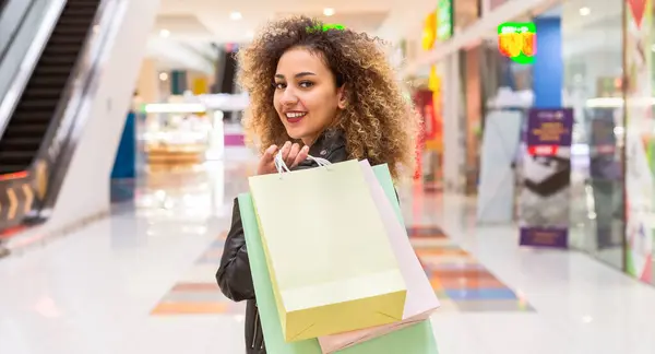 stock image A young African American woman is seen carrying multiple shopping bags in a crowded mall. She looks focused as she navigates through the bustling environment, surrounded by various stores.