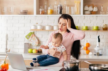 Bir anne bebeğini modern bir mutfakta kucağında tutarken uzaktan kumandalı işini yönetmeye çalışır. Ev işleri ve çocuk bakımı sorumluluklarının zorluklarını vurgular..