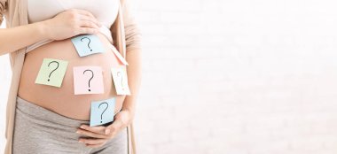 Hamile bir kadın göbeğine nazikçe dokunur. Üzerinde soru işaretleri olan renkli notlar vardır. Bu da bebeğin cinsiyeti hakkındaki merakı sembolize eder. Arka plan basit ve kapalı, kırpılmış, kopyalama alanı