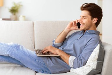 Bir adam, dizüstü bilgisayar kullanırken, bacakları yukarıda, koltukta gevşeyip telefonla konuşur..