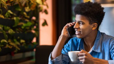 Afro-Amerikalı bir adam önünde bir fincan kahveyle bir masada oturuyor ve bir telefon görüşmesi yapıyor. Çağrıya odaklanmış ve özenli görünüyor..
