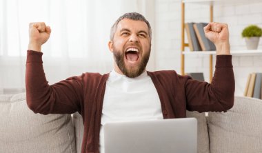Bir adam dizüstü bilgisayarla kanepede otururken görülüyor ve kolları havada. Enerjik görünüyor ve muhtemelen bir şeyleri kutluyor. Arka plan basit bir oturma odası..