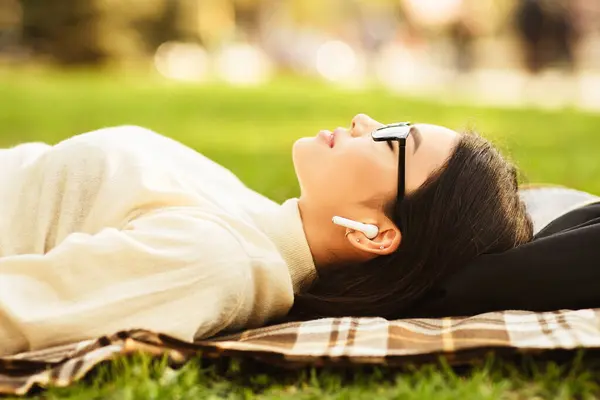 stock image Asian woman wearing sunglasses and earphones is lying on a plaid blanket in a sunny park. The woman appears relaxed, enjoying some leisure time on a warm afternoon, surrounded by greenery.