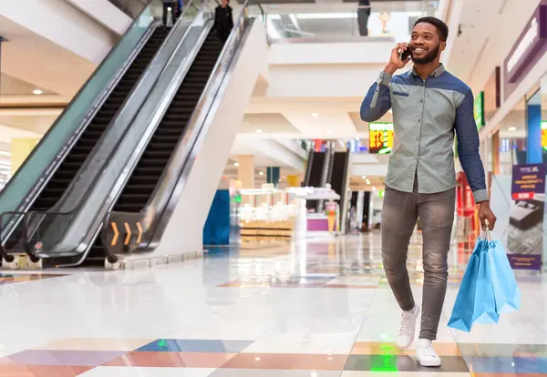 stock image A black man is seen walking through a bustling shopping mall, engaged in a phone conversation. He appears focused on his call amidst the hustle and bustle of the malls activity, copy space