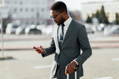Takım elbiseli Afro-Amerikalı bir adam havaalanı girişinin yanında bavuluyla dururken telefonunu kullanıyor. İş seyahatlerinin vurgulandığı modern ve profesyonel bir ortam..
