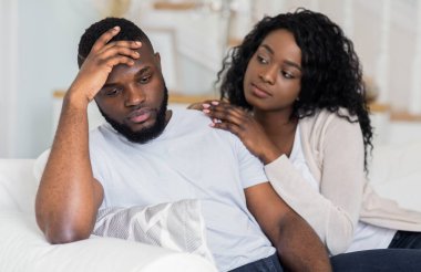 Bir kadın kanepede oturan bir adamın yanına oturur, eli omzunda otururken o da başını kaldırıp aşağıya bakar..