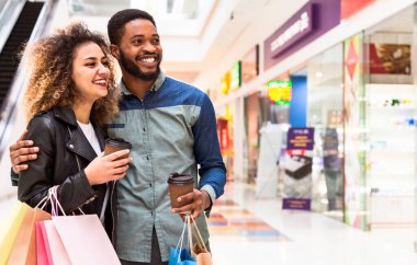 Neşeli siyahi bir erkek ve kadın alışveriş merkezlerinde ellerinde kahve poşetleri, sohbet ve kahkahalar ile dolaşır, günlük eğlence ve perakende terapi anlarını sergilerler..