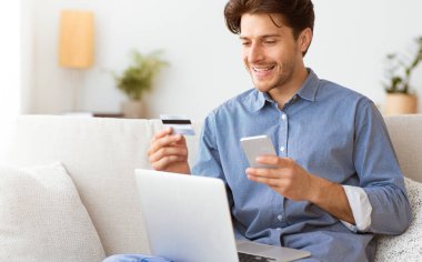 Kanepede oturan bir adam, internette kredi kartı ve akıllı telefon kullanarak ödeme yaparken gülümsüyor..
