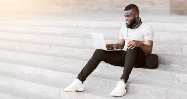 Şehir ortamında beton merdivenlerde oturan bir adam dizüstü bilgisayarla çalışırken gülümsüyor..