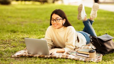 Asyalı bir kadın önündeki dizüstü bilgisayarla çimlere uzanmış güneşli bir günde parkta yazı yazıyor ve çalışıyor..