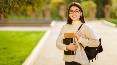 Gözlüklü Çinli bir kadın bir elinde kitap, diğer elinde sırt çantası tutarken ayakta duruyor..