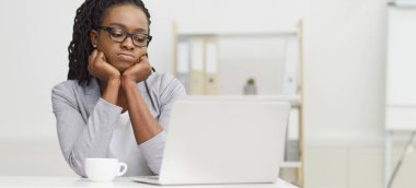 Yorgun siyah bir kadın ofis masasında dizüstü bilgisayarını kullanıyor. Karmaşık bir masa ve sönük bir çevreyle, sıkıcı ve yorgun bir ruh hali içinde, işiyle ilgilenmediğini gösteriyor..