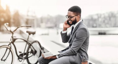 Profesyonel siyahi adam dizüstü bilgisayarını kullanıyor ve yanında bir bisikletle dışarıda otururken telefonla konuşuyor. Sahne modern ve enerjik, yeni bir ortamda çoklu görev..