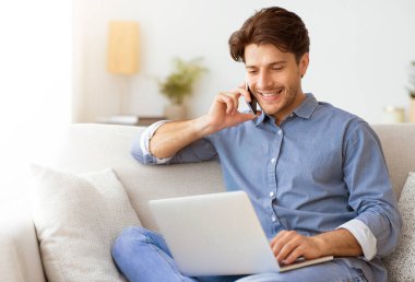 Bir adam kanepede oturuyor, telefonuyla konuşurken gülümsüyor ve laptopuyla çalışıyor..
