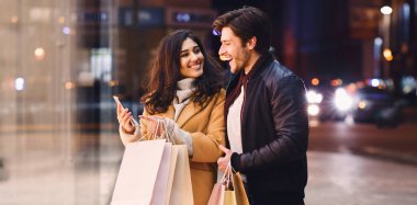 Bir adam ve bir kadın işlek bir caddede ellerinde alışveriş torbalarıyla yürüyorlar..