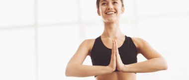 Siyah spor sutyen giyen bir kadın, elleri Namaste pozunda yoga yaparken, parlak bir stüdyoda, fotokopi alanı olan bir panoramada gülümserken görülüyor.