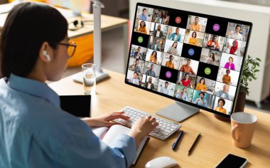 Kulaklık takan bir kadın, birçok katılımcıyla bir video konferans görüşmesi yapan bir bilgisayar monitörünün önünde masasında oturuyor. Toplantıya aktif olarak katılıyor.