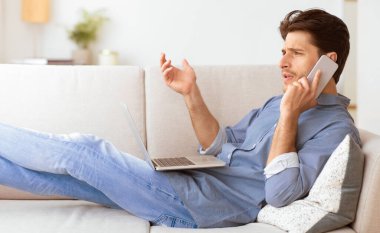 Kanepede oturan bir adam dizüstü bilgisayarı üzerinde çalışırken telefonla konuşuyor..