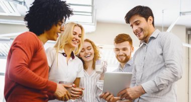 Farklı etnik gruplardan oluşan bir grup, bir tablet cihazın etrafında toplanmış, ekranda gösterilen içeriğe odaklanmış, bir proje üzerinde tartışıyor veya işbirliği yapıyor..